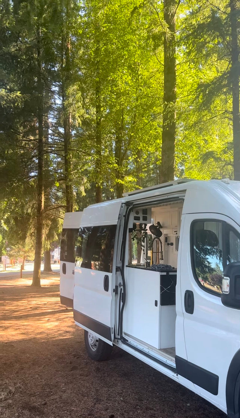 A bright white RV rental with its side door open, revealing a modern custom interior kitchen, parked among tall trees for camping. — photo 13