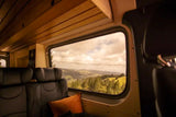 Stunning views from an RV rental's cozy interior. Lush green hills & forests seen through a large window, under a cloudy sky.