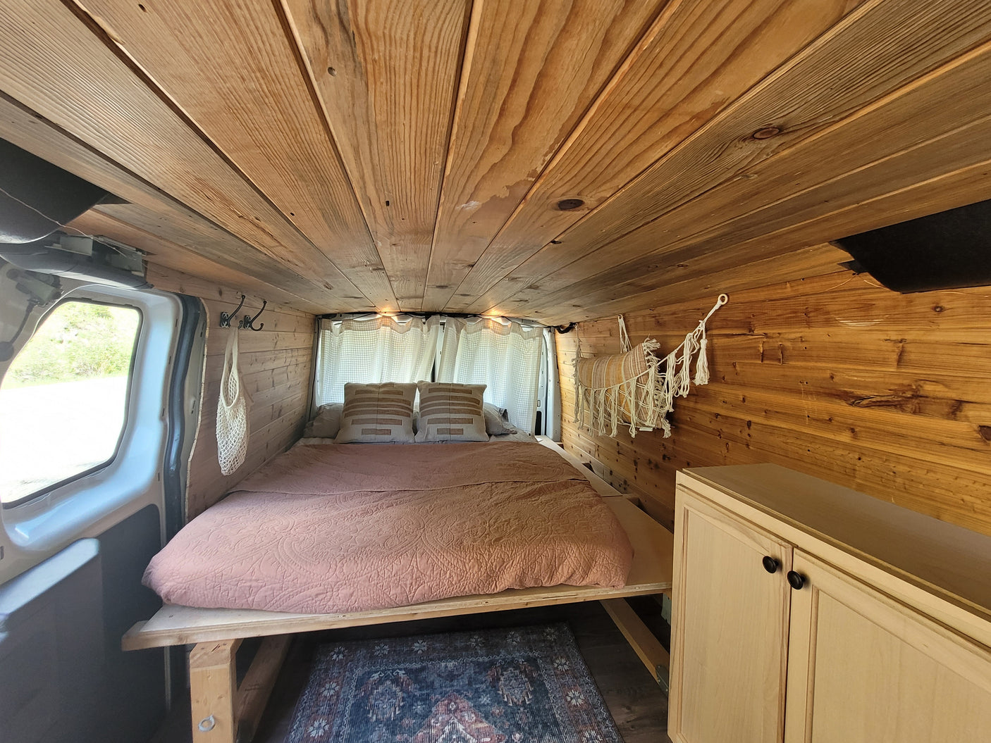 Cozy, wood-paneled RV rental interior: bed with pillows & comforter, hanging storage, and cabinet. Perfect for van life.