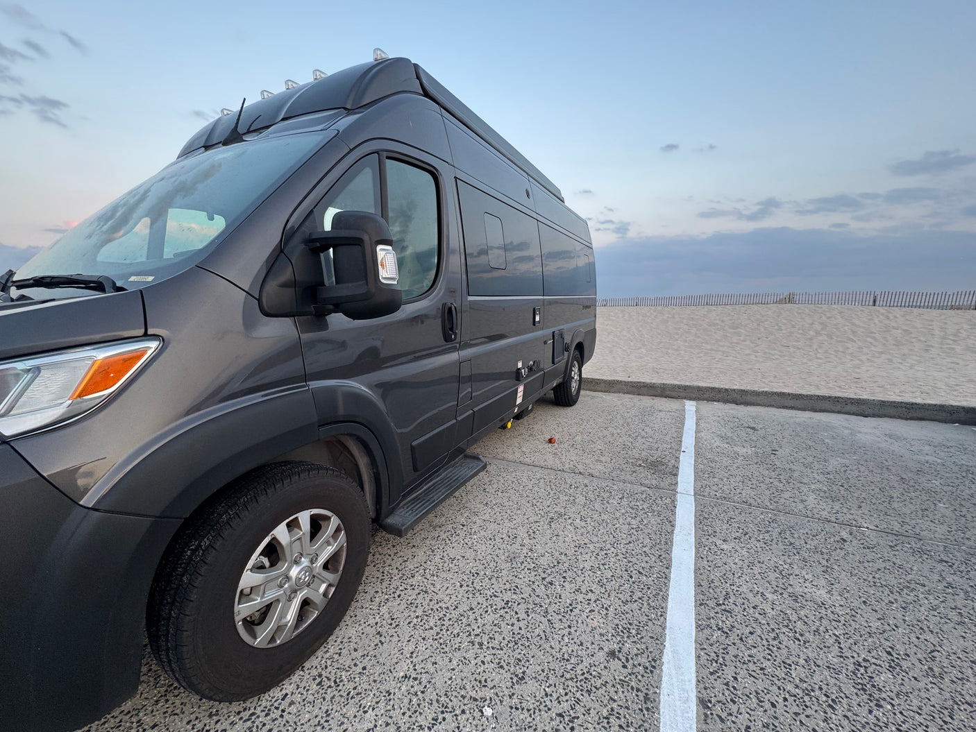 Dark gray RV rental parked on a speckled lot, overlooking a sandy beach with a fence under a cloudy sky.