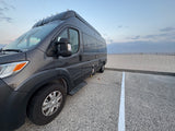 Dark gray RV rental parked on a speckled lot, overlooking a sandy beach with a fence under a cloudy sky.