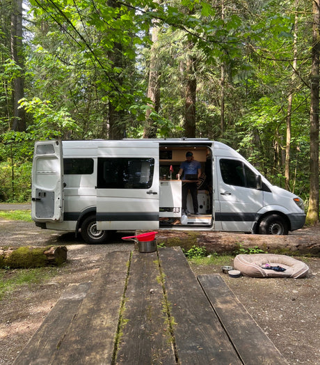Man cooking inside a white RV rental van at a forest campsite, with a picnic table and dog bed nearby.