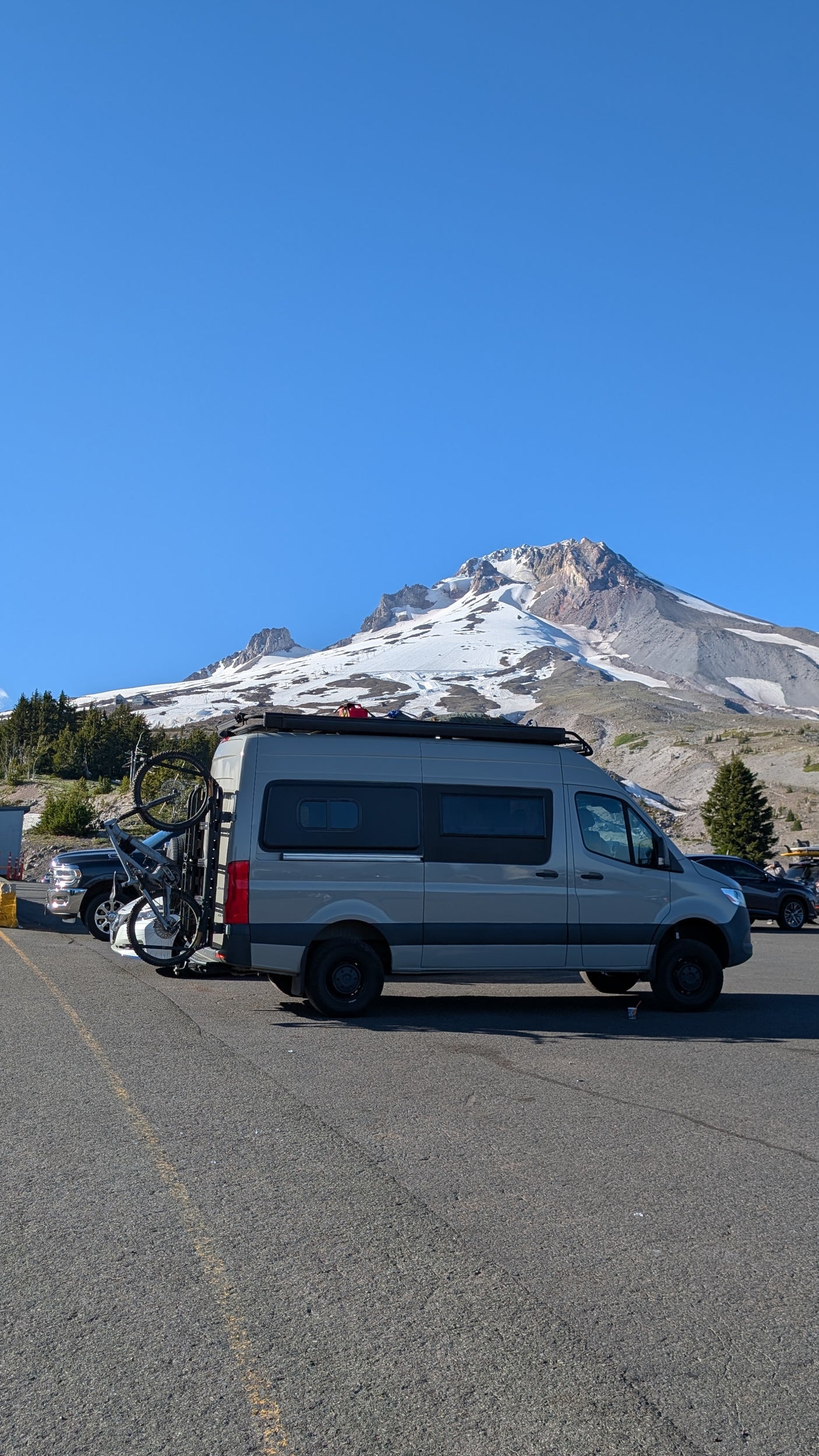 This gray camper van, perfect for an RV rental adventure, is parked with a bike rack against a stunning snow-capped mountain.