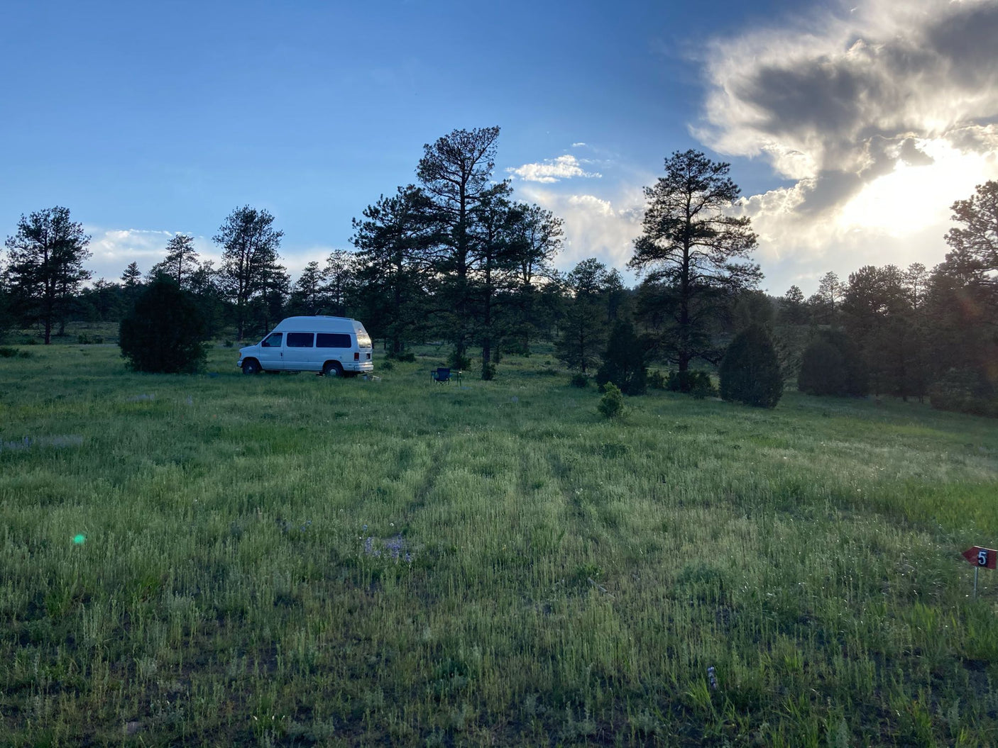 Scenic rv rental campsite featuring a white camper van, chairs, and trees under a dramatic cloudy sky, perfect for outdoor adventure.