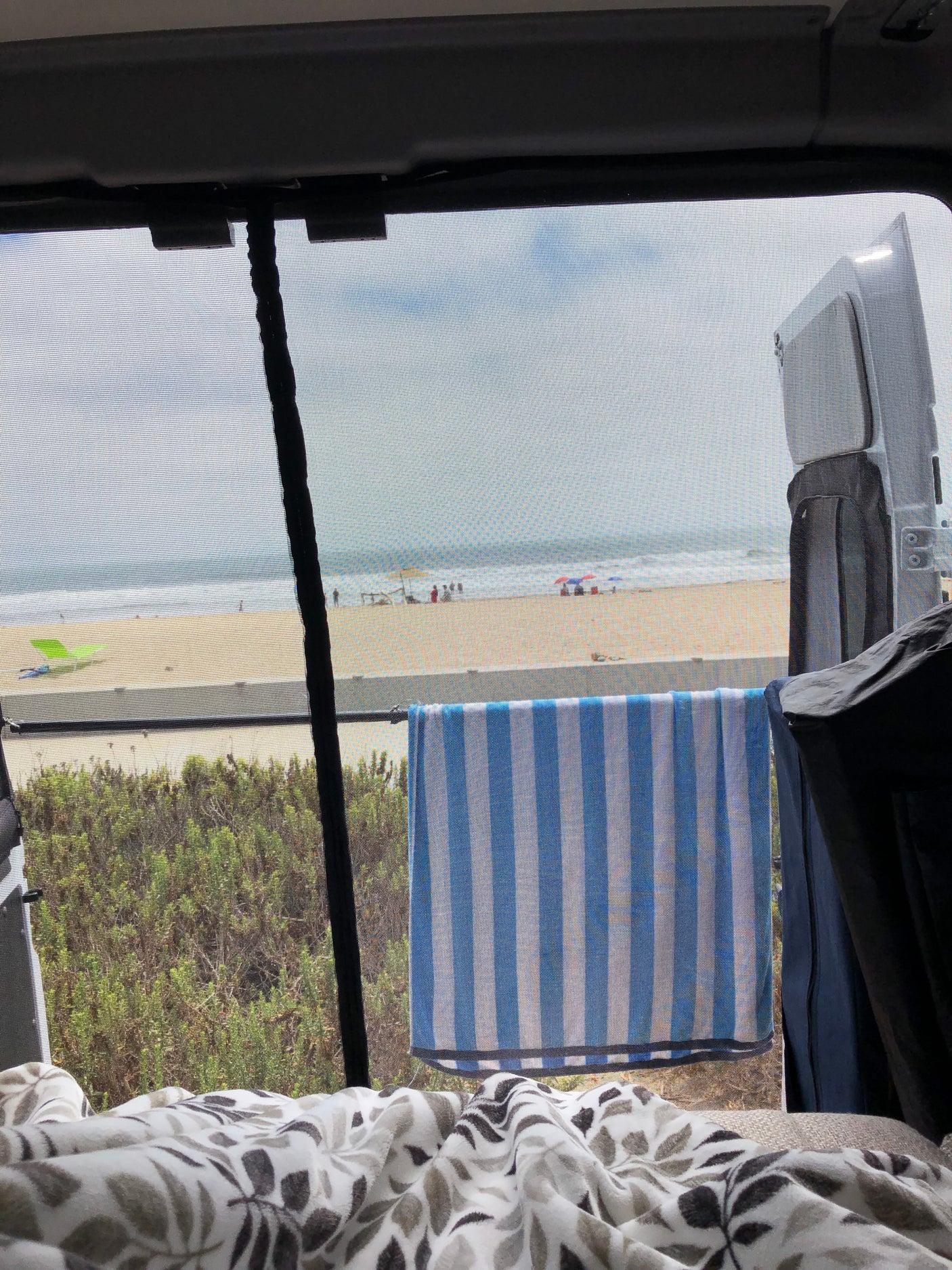 Cozy RV rental view: ocean waves, sandy beach, and lush green. Screened window, striped towel, comfy blanket.