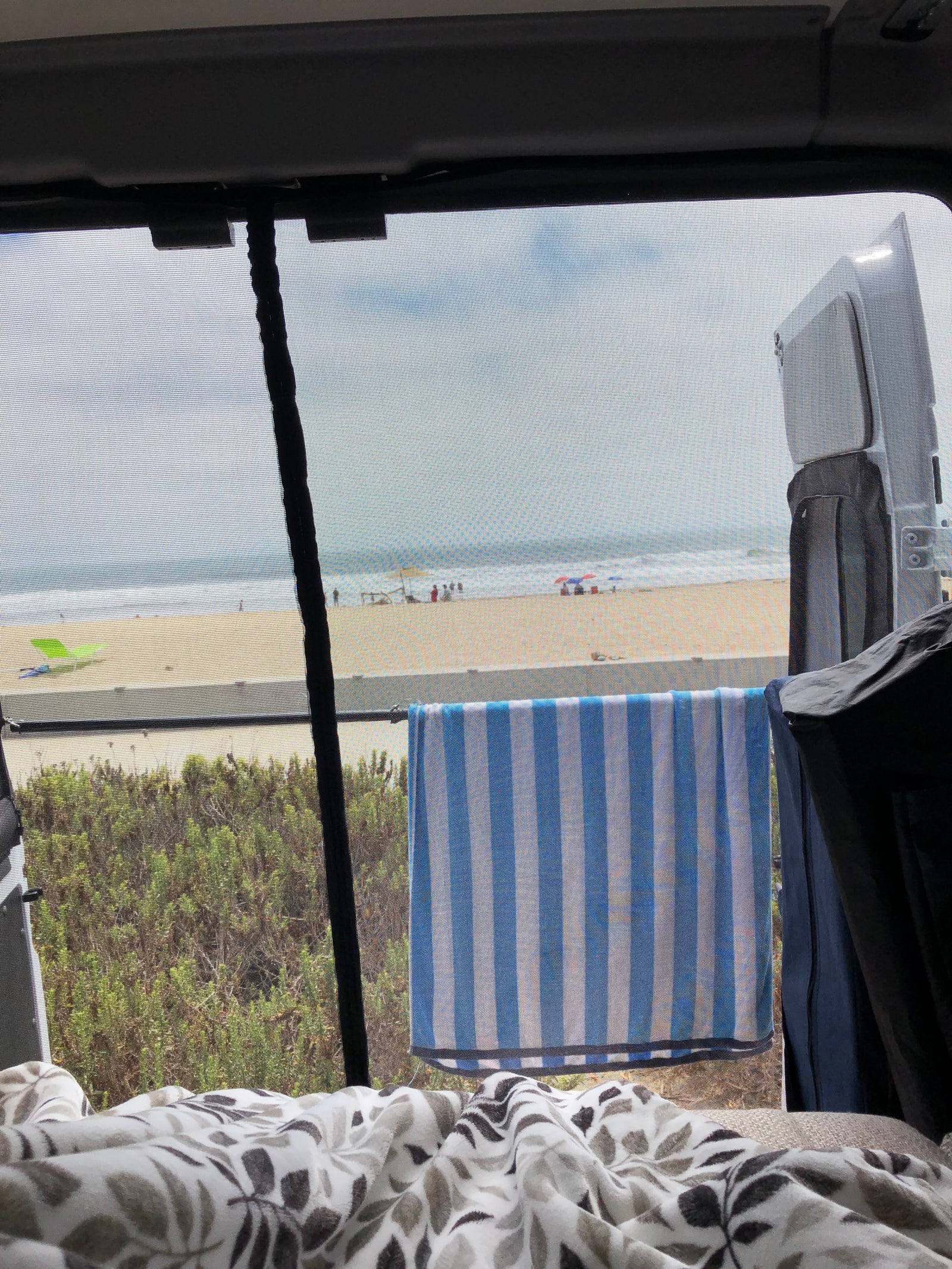 Cozy RV rental view: ocean waves, sandy beach, and lush green. Screened window, striped towel, comfy blanket.