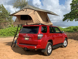 A red Toyota 4Runner with a tan rooftop tent and ladder, parked on a dirt path overlooking the ocean. Great RV rental for beach camping.