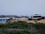 A white RV rental van and SUV parked on a misty, rocky coast overlooking the ocean with waves and a bench.