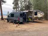 RV rental: Gray camper van with bikes and large fifth-wheel RV parked at a lakeside forest campsite, ready for adventure.