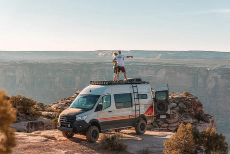 This custom Mercedes Sprinter van, perfect for an RV rental adventure, is parked on a cliff. People admire the canyon view from its roof.