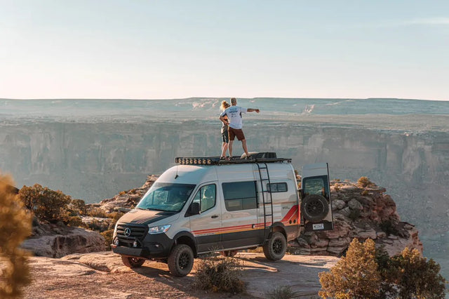 This custom Mercedes Sprinter van, perfect for an RV rental adventure, is parked on a cliff. People admire the canyon view from its roof.