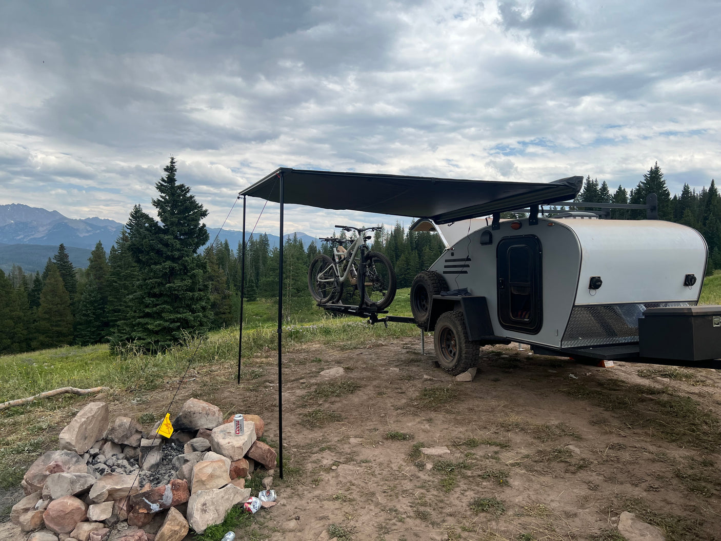 Scenic RV rental campsite: A teardrop trailer with bikes, an awning, and a fire pit, surrounded by pine trees and mountains under cloudy skies.