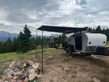 Scenic RV rental campsite: A teardrop trailer with bikes, an awning, and a fire pit, surrounded by pine trees and mountains under cloudy skies.