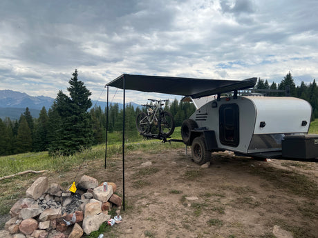 Scenic RV rental campsite: A teardrop trailer with bikes, an awning, and a fire pit, surrounded by pine trees and mountains under cloudy skies.
