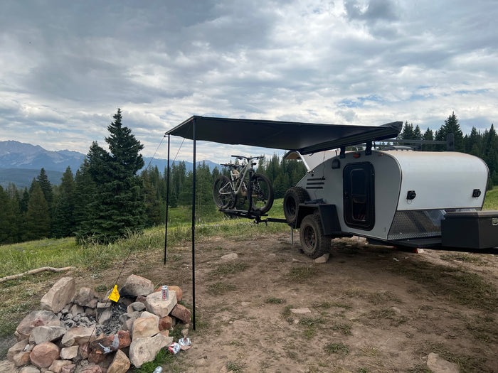 Scenic RV rental campsite: A teardrop trailer with bikes, an awning, and a fire pit, surrounded by pine trees and mountains under cloudy skies.
