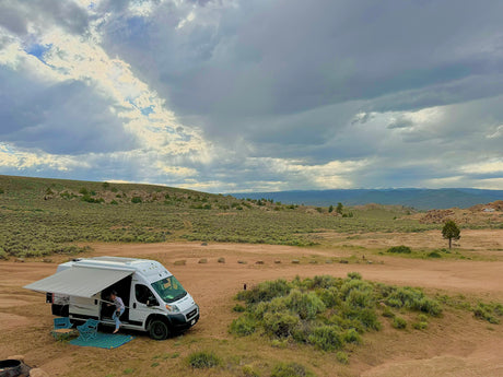 A white RV rental camper van, awning extended, is parked in a vast, arid landscape as a person steps out under a cloudy sky.
