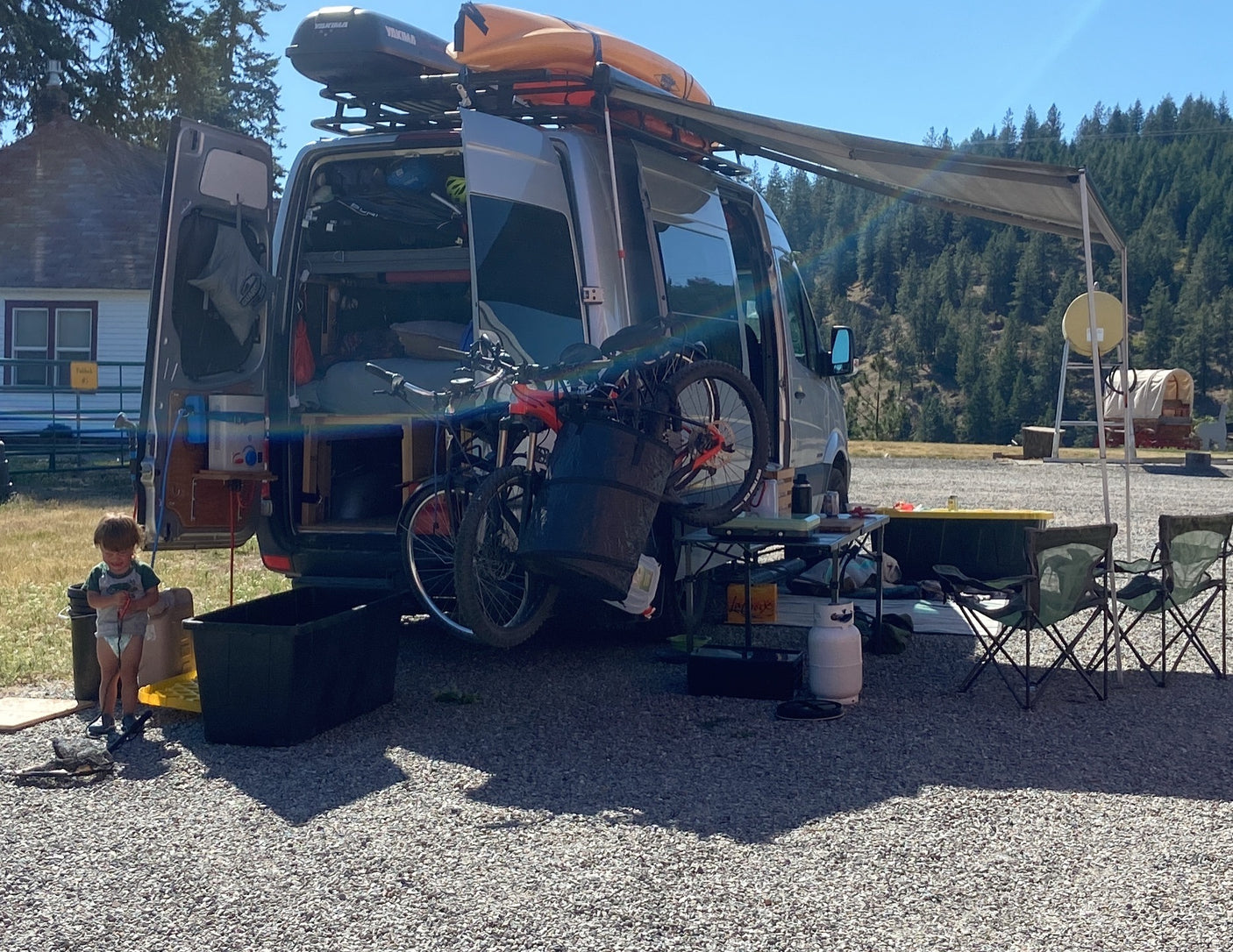 Camper van RV rental set up with bikes, kayak, and awning. A child plays near the open van, perfect for outdoor family travel.