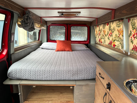 An RV rental's clever interior design shows a comfortable bed, floral curtains, and a compact kitchen counter with sink.