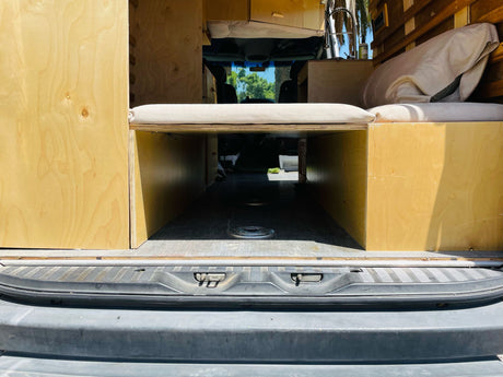 Inside an RV rental camper van: light wood storage, bed platform with cushions, looking forward through the windshield.