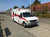 White ambulance-style RV rental with red stripe details, parked on a sunny street by coastal homes.
