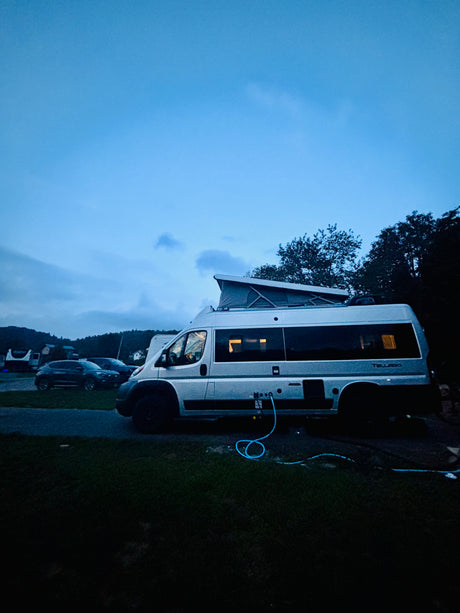 Night scene showing a silver rv rental campervan with a pop-top roof at a campsite, hooked up to utilities. Lights glow inside.