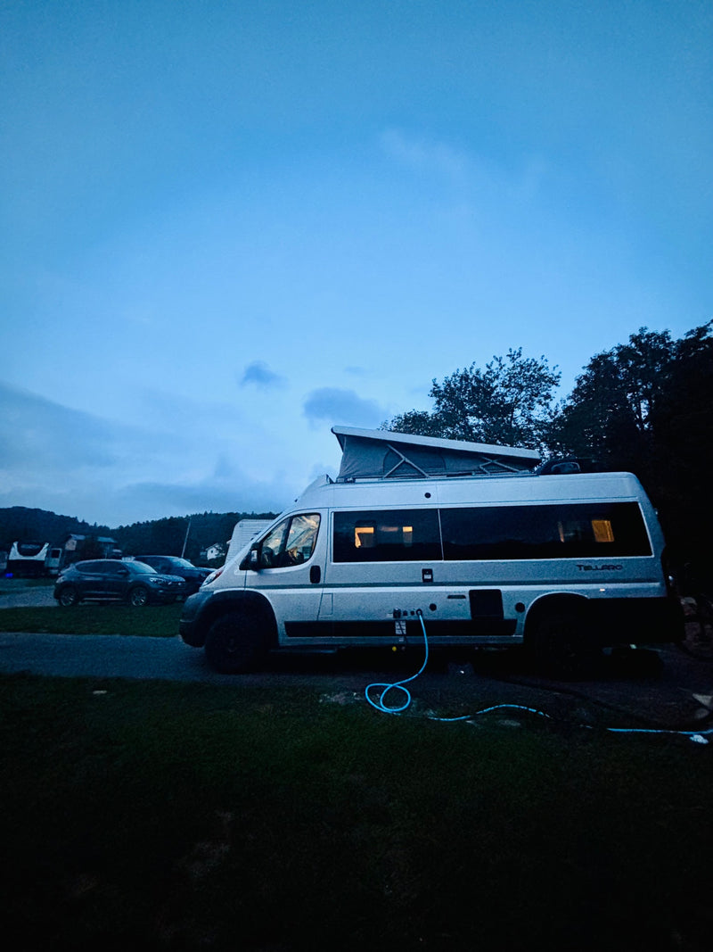Night scene showing a silver rv rental campervan with a pop-top roof at a campsite, hooked up to utilities. Lights glow inside. — photo 10
