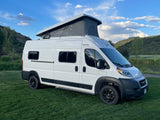 White pop-top camper van, an RV rental, parked on green grass with scenic mountains and a calm pond nearby.