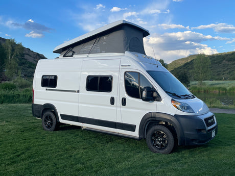 White pop-top camper van, an RV rental, parked on green grass with scenic mountains and a calm pond nearby.