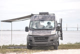 Explore with this dark grey Ram ProMaster camper van. Awning deployed by water with bridge view. Ideal rv rental for adventure!