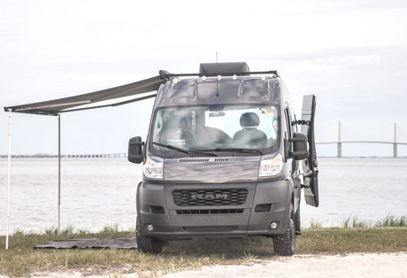 Explore with this dark grey Ram ProMaster camper van. Awning deployed by water with bridge view. Ideal rv rental for adventure!