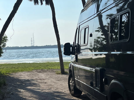 Enjoying nature in an RV rental: a dark camper van parked by a serene lake, framed by palm trees.
