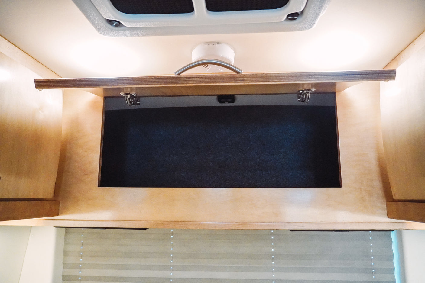 Open wooden cabinet storage in an RV rental, showing ceiling vent, smoke detector, and window blinds.