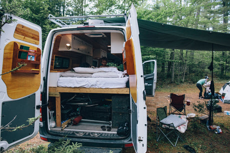 Experience forest camping with an RV rental! A child is cozy inside the custom camper van, while an adult sets up camp outside.