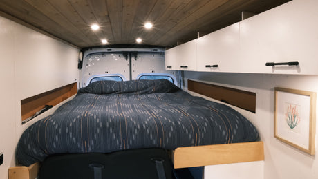 RV rental interior: cozy camper van bedroom with patterned duvet, overhead cabinets, and a wood-paneled ceiling.