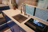 Modern RV rental kitchen featuring sleek white counters, a sink, and induction cooktop. Blue cabinets brighten the space.