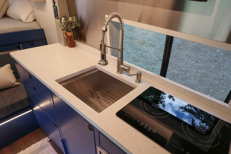 Modern RV rental kitchen featuring sleek white counters, a sink, and induction cooktop. Blue cabinets brighten the space.