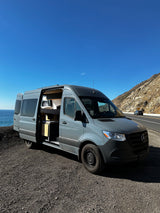 Grey RV rental camper van by ocean on a coastal highway, side door open showing interior. Ideal for scenic travel & van life.