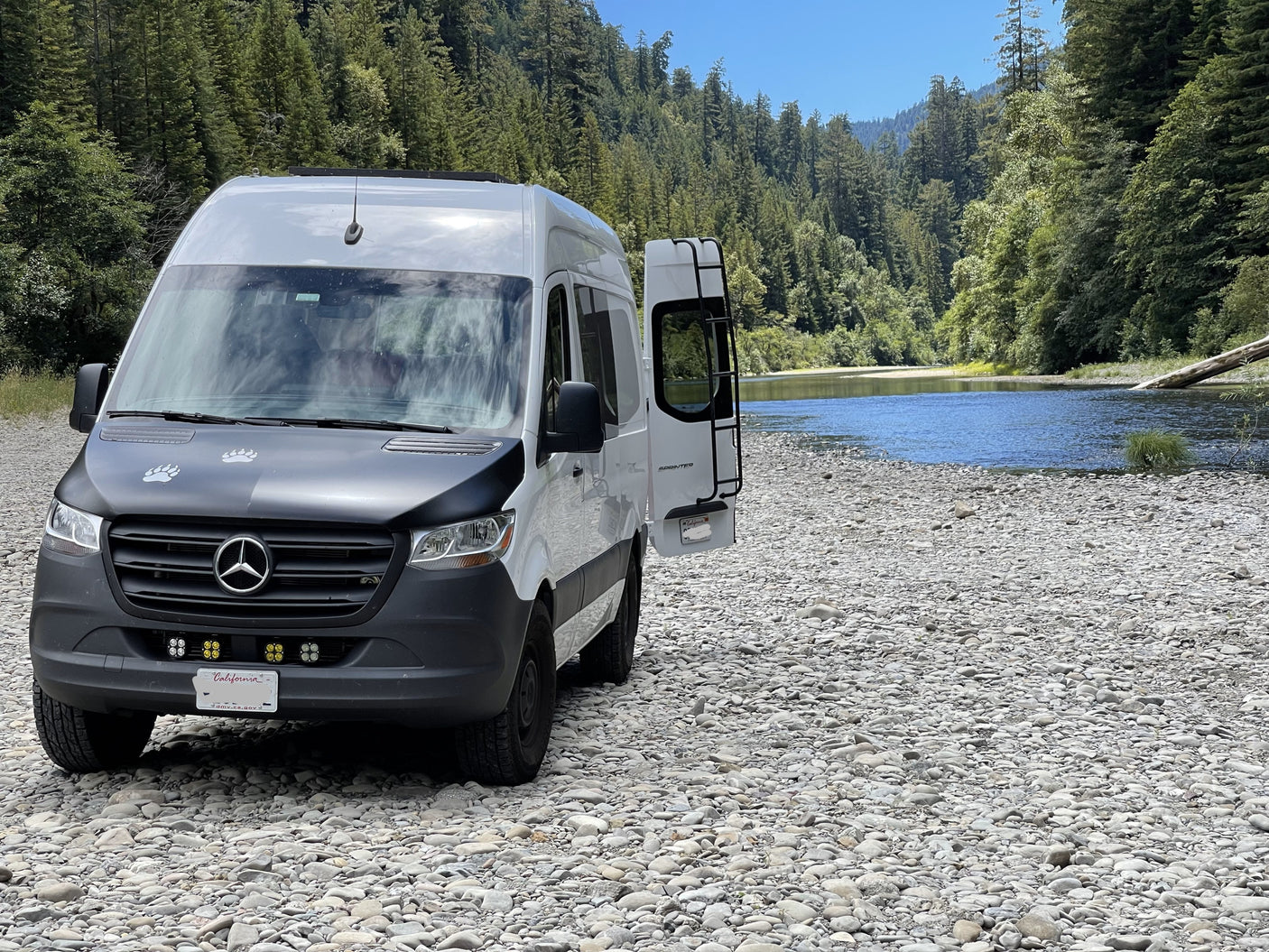 RV rental: Mercedes Sprinter camper van on a rocky riverbank by a blue river, framed by forested mountains.