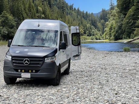 RV rental: Mercedes Sprinter camper van on a rocky riverbank by a blue river, framed by forested mountains.