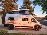 A white Winnebago Solis camper van with a raised pop-top roof, bathed in golden sunset light on a driveway, ideal for an RV rental.