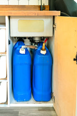 RV rental: Under-sink setup with two blue tanks for freshwater and greywater storage. Compact water system.
