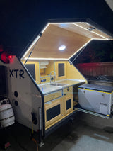 Compact RV rental teardrop camper's illuminated outdoor kitchen with sink, storage, and pull-out fridge for adventures.