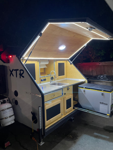 Compact RV rental teardrop camper's illuminated outdoor kitchen with sink, storage, and pull-out fridge for adventures.