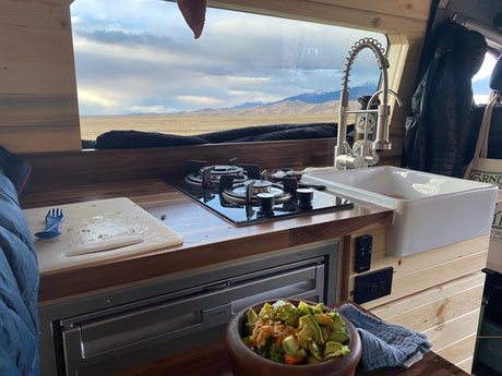 A modern RV rental kitchen with a stove, sink, and fridge, offering a stunning view of desert dunes and distant mountains. Salad on counter.