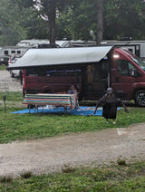 Rainy campground scene: a red RV rental with an awning provides shelter. A person is seated at a striped table, another in a poncho.