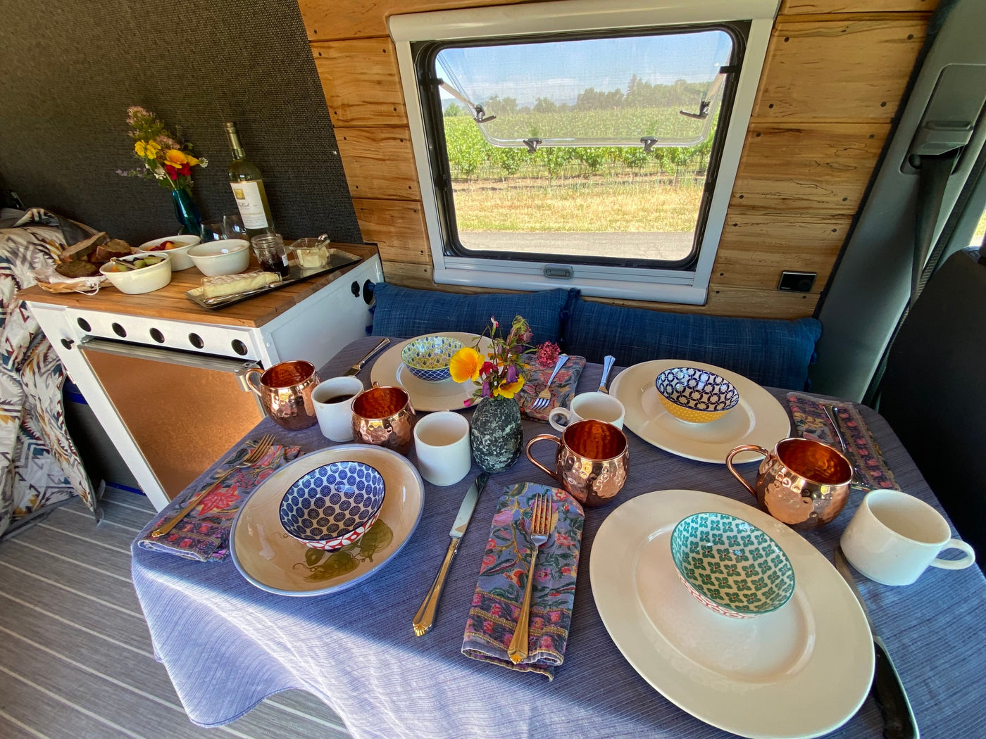 A cozy RV rental interior with a table set for a meal, featuring fruit, cheese, wine, and a scenic vineyard view outside. — photo 7