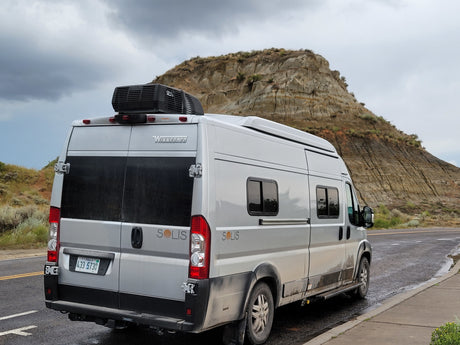 Silver Winnebago Solis rv rental camper van on a wet road, backed by a layered rock formation under cloudy skies.