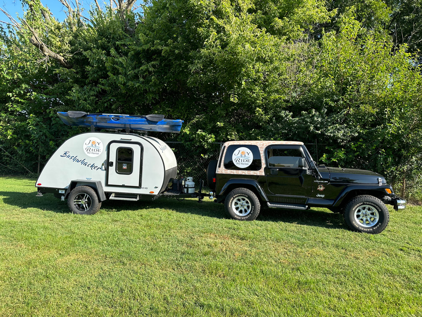 Black Jeep and white RV rental with kayak parked on green grass by trees, displaying "Joy Ride RV Rental" logos.