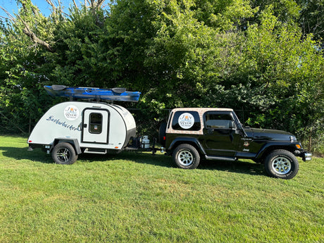 Black Jeep and white RV rental with kayak parked on green grass by trees, displaying "Joy Ride RV Rental" logos.