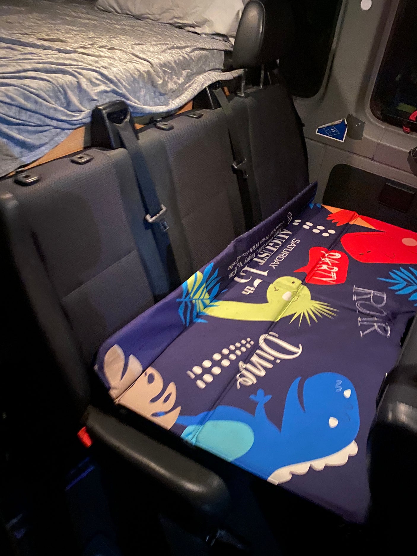 RV rental camper van interior featuring a cozy bed, passenger seats, and a fun dinosaur mat for kids.